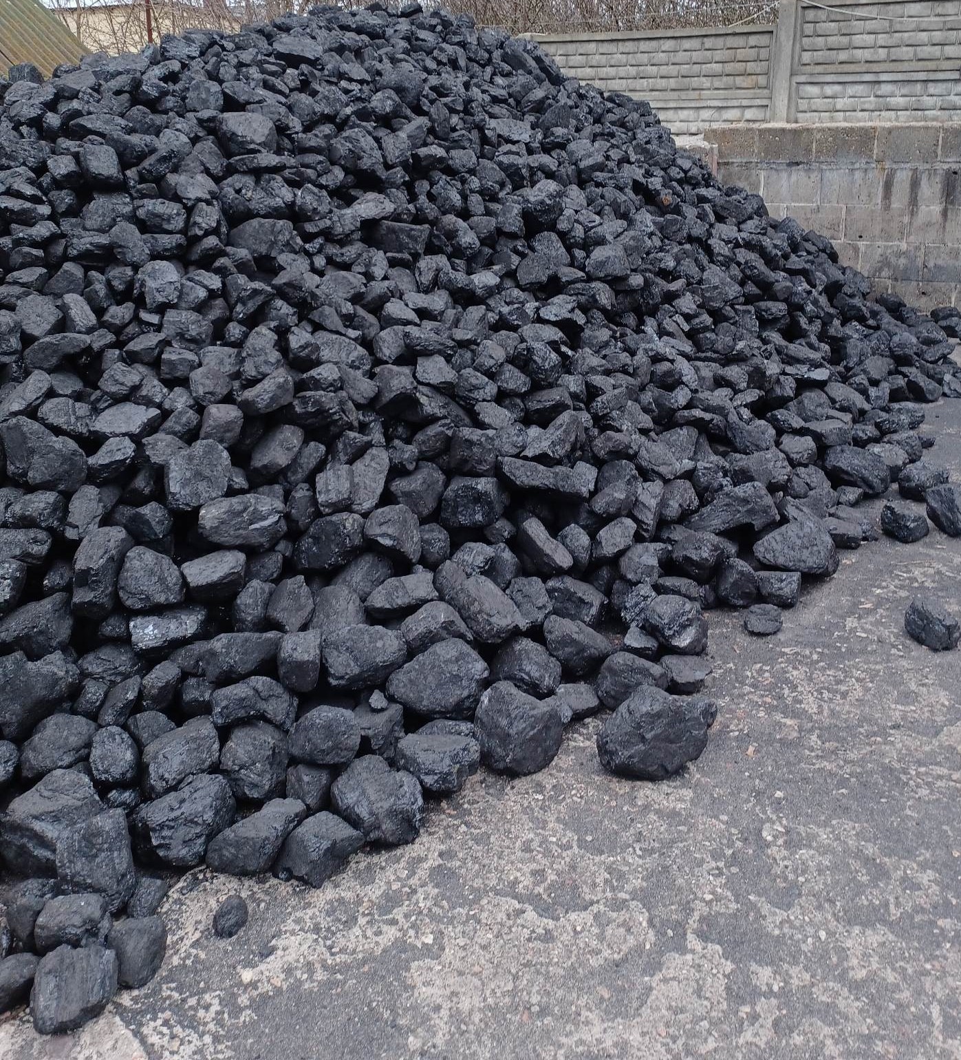 Góra czarnego węgla kamiennego na betonowym podłożu, widoczny fragment betonowego muru w tle.
