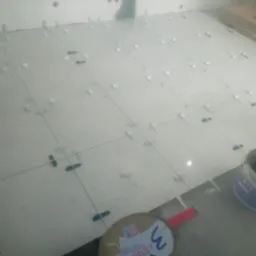 Układanie białych płytek podłogowych z użyciem krzyżyk&oacute;w dystansowych, widoczny pojemnik z zaprawą i poziomica.