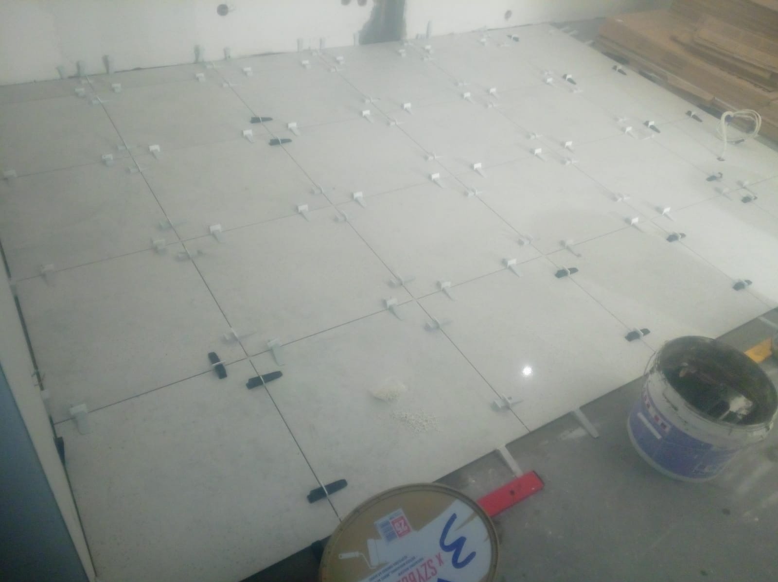 Układanie białych płytek podłogowych z użyciem krzyżyków dystansowych, widoczny pojemnik z zaprawą i poziomica.