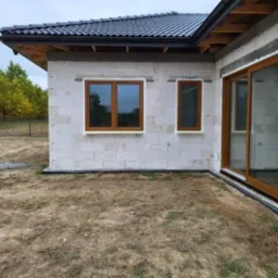 Surowy stan budynku w trakcie budowy z widocznymi oknami z brązowymi ramami oraz przesuwnymi drzwiami tarasowymi, ściany z białego bloczka, teren wokół niezagospodarowany.