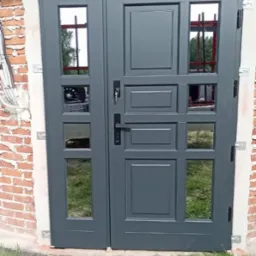 Szare, dwuskrzydłowe drzwi wejściowe z przeszkleniami, zamontowane w murze z czerwonej cegły, widoczne elementy montażowe.