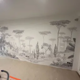 Ściana po tapetowaniu z motywem krajobrazu toskańskiego w odcieniach szarości, widoczne gniazdka elektryczne i fragment podłogi w trakcie remontu.