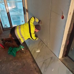 Euroserwis Serhii Zalevskyi - Pracownik w odblaskowej kurtce układa szare płytki imitujące marmur na podłodze w surowym wnętrzu, widoczne narzędzia i poziomica.