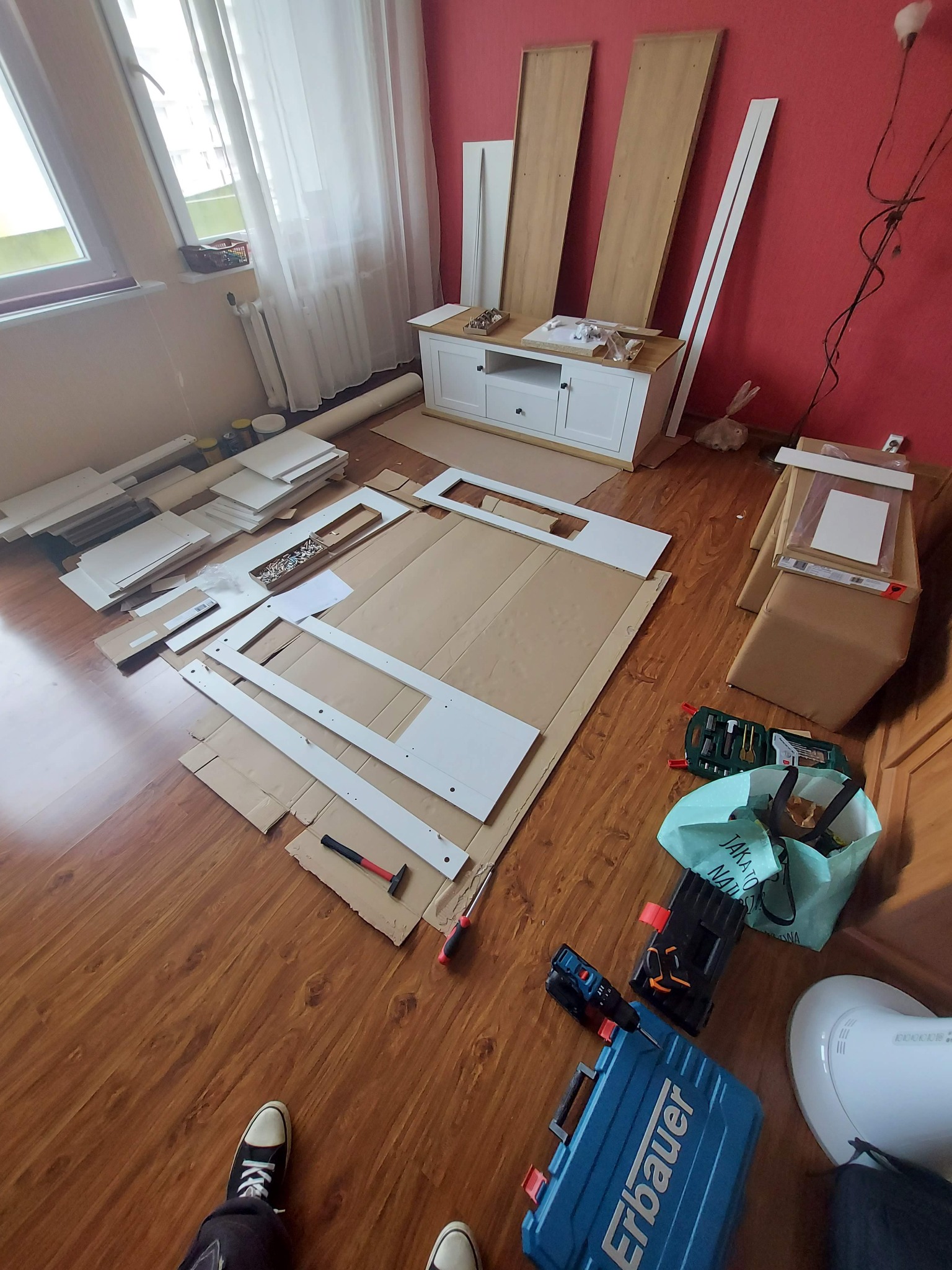 Rozłożone elementy białej komody na podłodze z paneli, narzędzia montażowe w pobliżu, widoczne skrzynki z narzędziami Erbauer, kartony zabezpieczające podłoże.