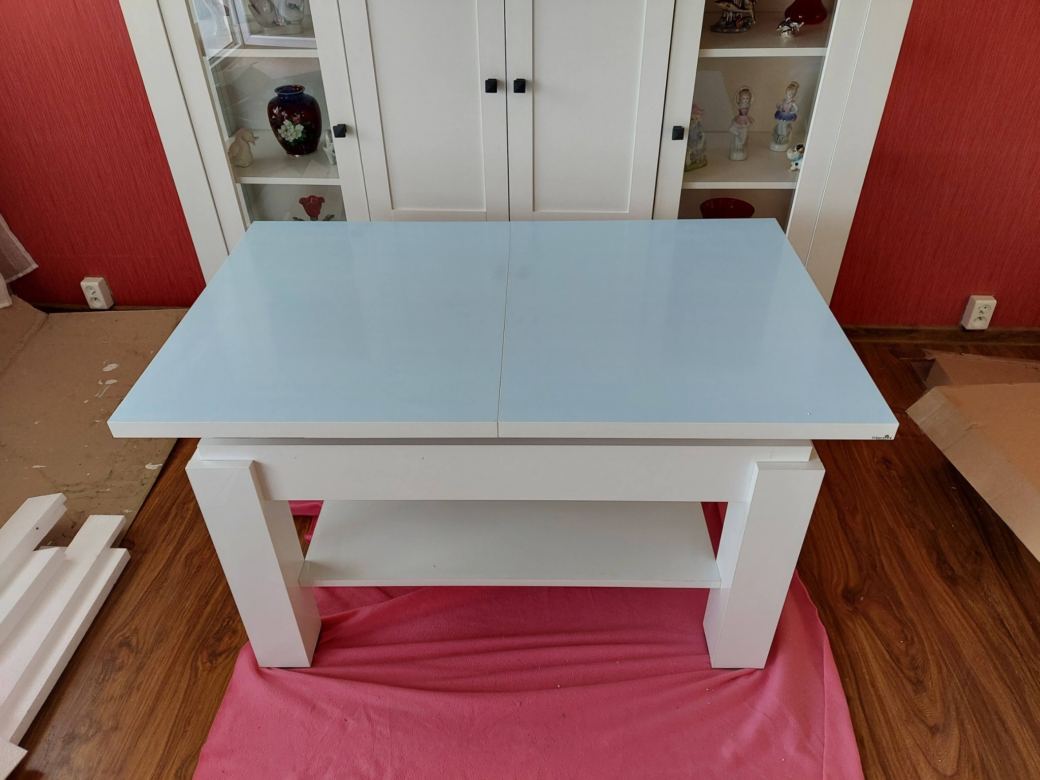 Biały stół z jasnoniebieskim blatem, częściowo rozłożony, na podłodze różowy materiał zabezpieczający, w tle biała szafka z ekspozycją i czerwona ściana z gniazdkiem.