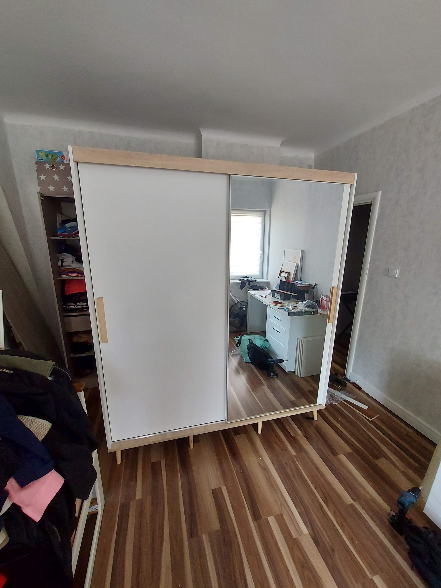 Nowoczesna szafa przesuwna z lustrem i białymi drzwiami, z drewnianymi akcentami, ustawiona na drewnianej podłodze w pokoju z widocznym biurkiem i narzędziami w odbiciu lustra.