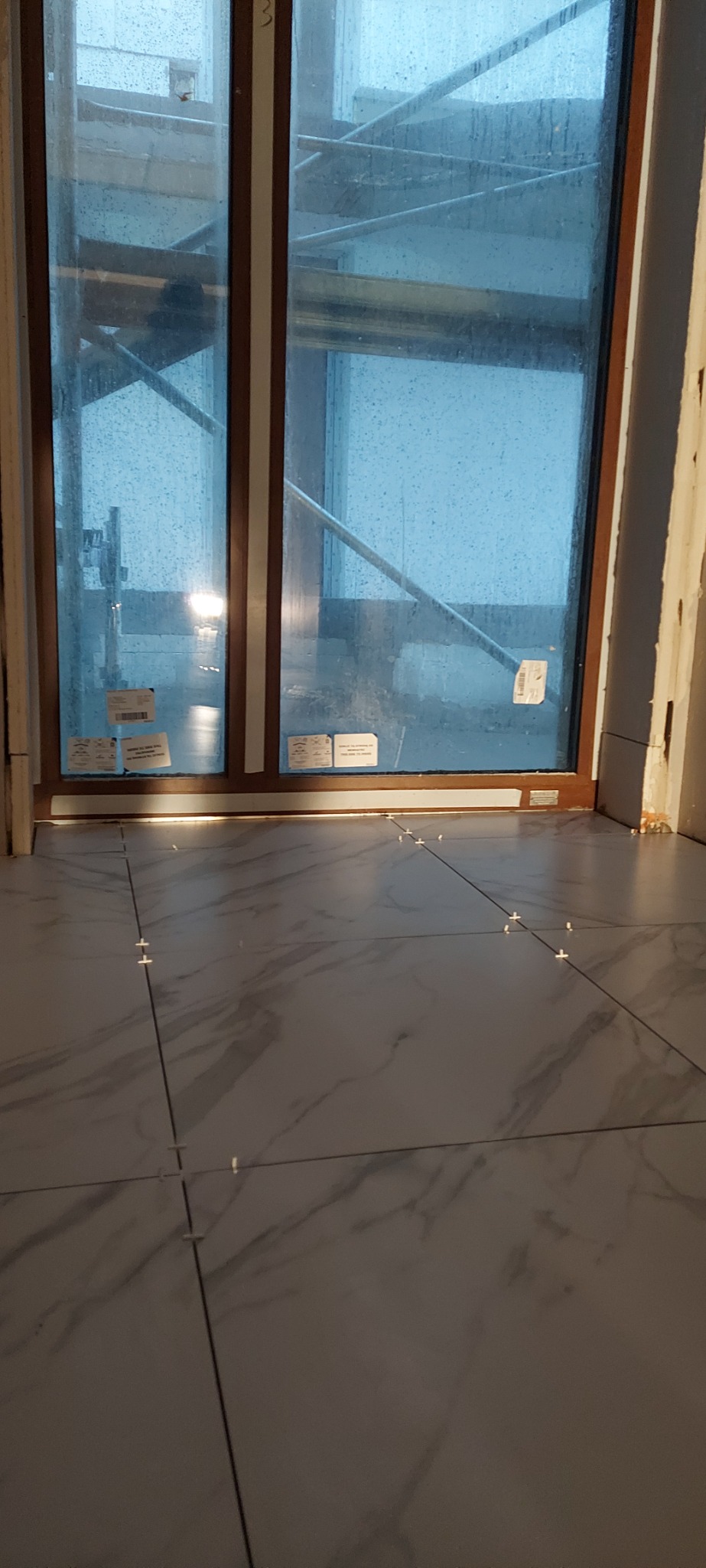 Podłoga wyłożona szarymi płytkami imitującymi marmur, z plastikowymi krzyżykami dystansowymi w spoinach, widok na okno z rusztowaniem na zewnątrz.