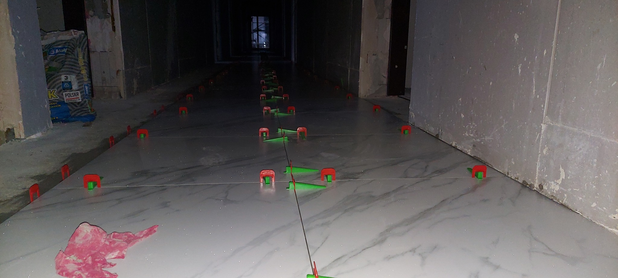 Układanie białych płytek imitujących marmur w długim korytarzu, z użyciem systemu poziomowania płytek (czerwono-zielone klipsy), widoczny worek z klejem do płytek w tle.