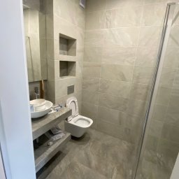 Nika-Bud - Nowoczesna łazienka w trakcie wykańczania, widoczne płytki imitujące kamień, zabudowa WC, umywalka nablatowa z pozostawionym wiadrem po farbie i narzędziami, kabina prysznicowa z przezroczystego...