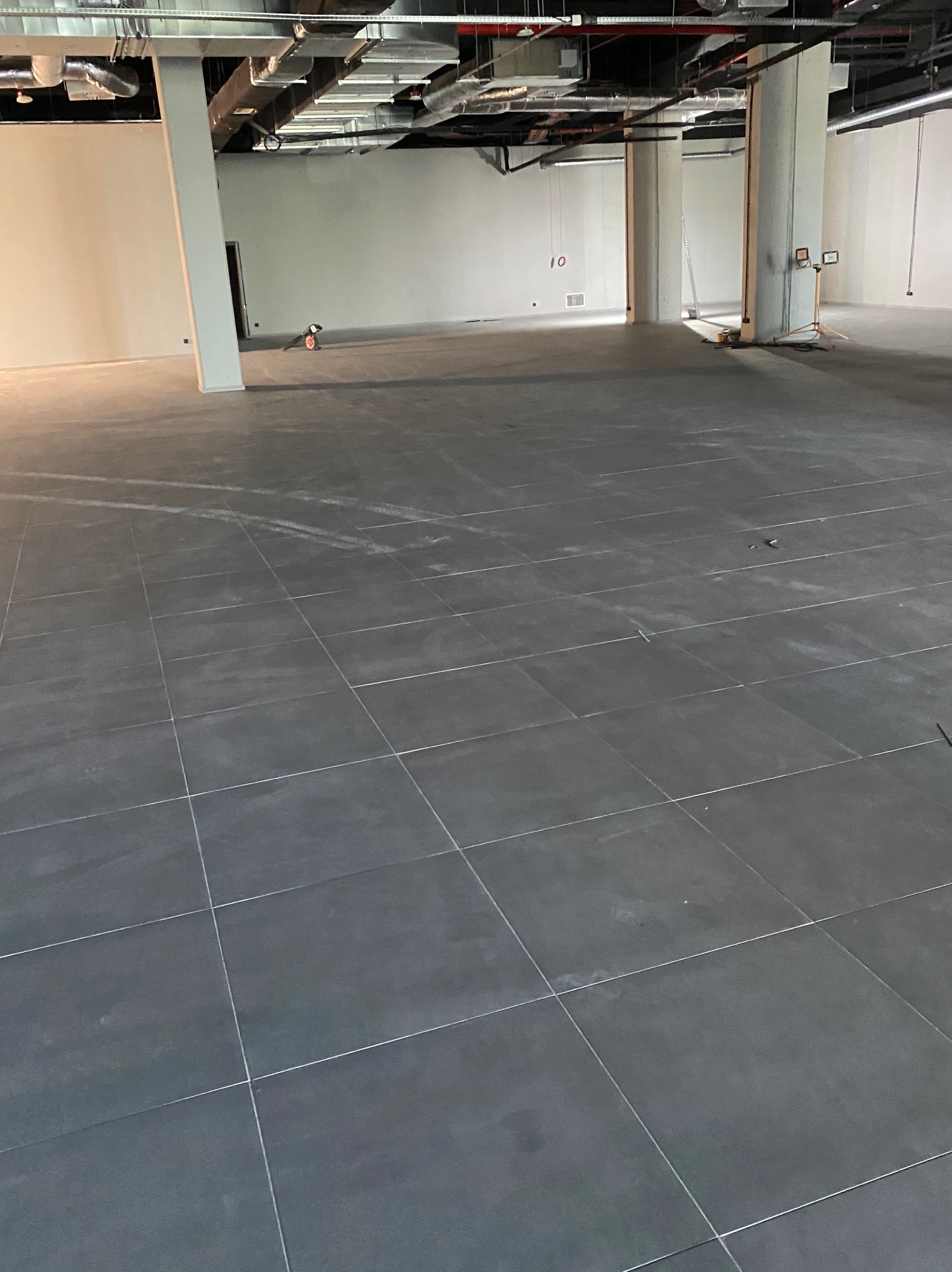 Duża powierzchnia wyłożona ciemnymi, kwadratowymi płytkami, z widocznymi śladami po budowie, oświetlona przez światło wpadające z zewnątrz, z widocznymi elementami instalacji pod sufitem...