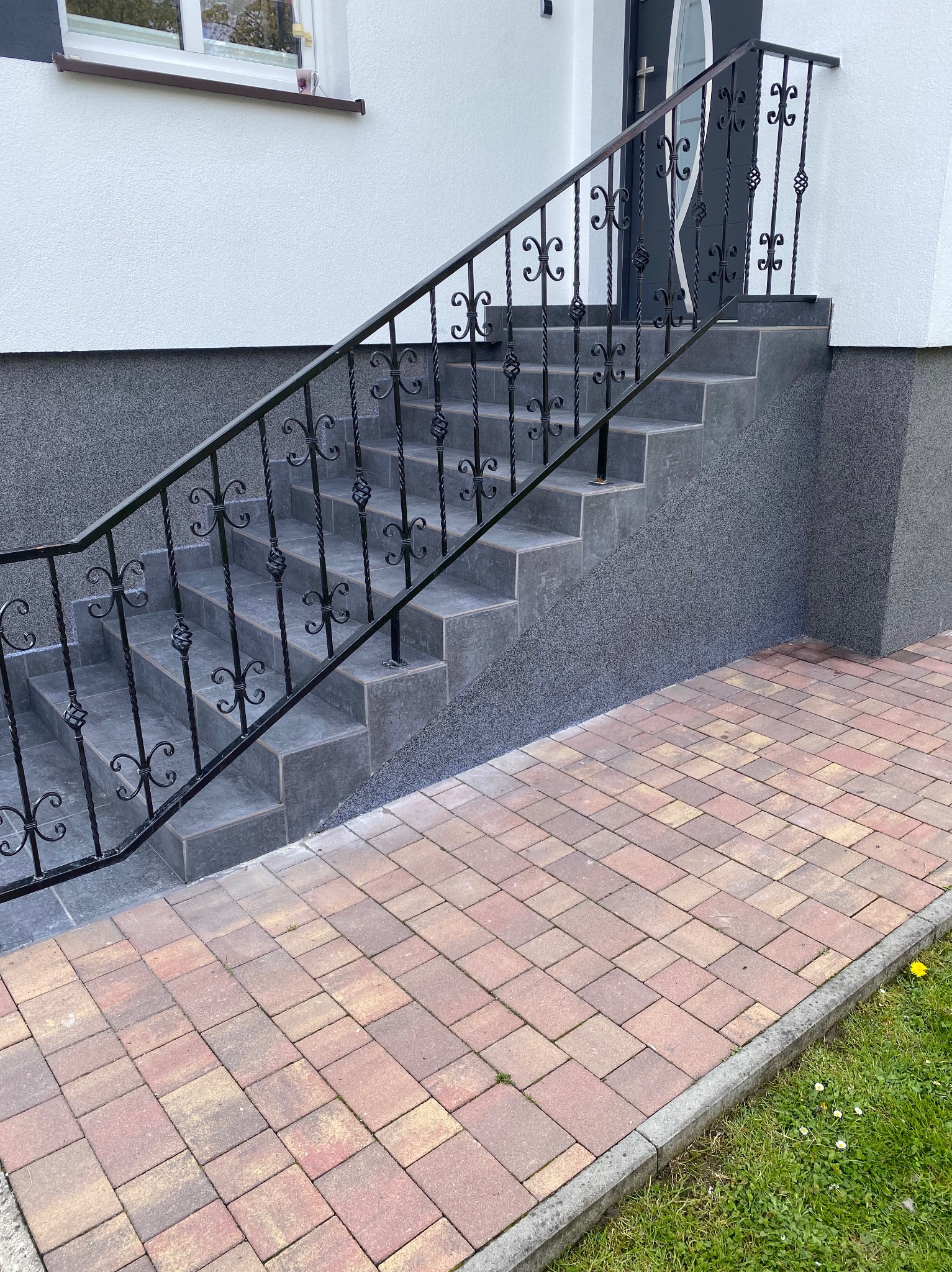 Zewnętrzne schody wejściowe wyłożone szarymi płytkami, z czarną, ozdobną balustradą, prowadzące do drzwi wejściowych, obok chodnik z kostki brukowej.