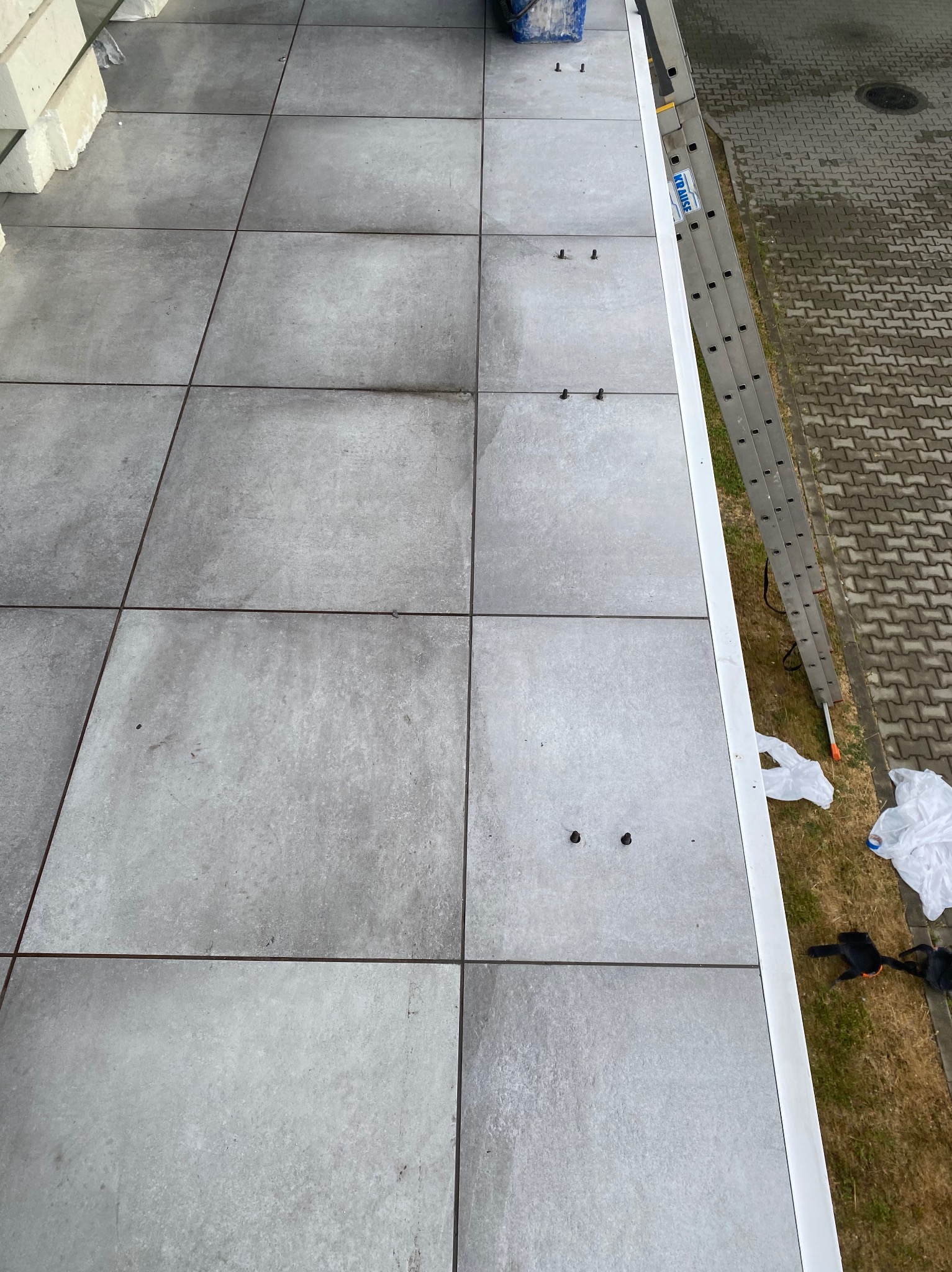 Ułożone szare płytki na tarasie z widocznymi szczelinami, drabina oparta o krawędź, przygotowane materiały budowlane w tle.