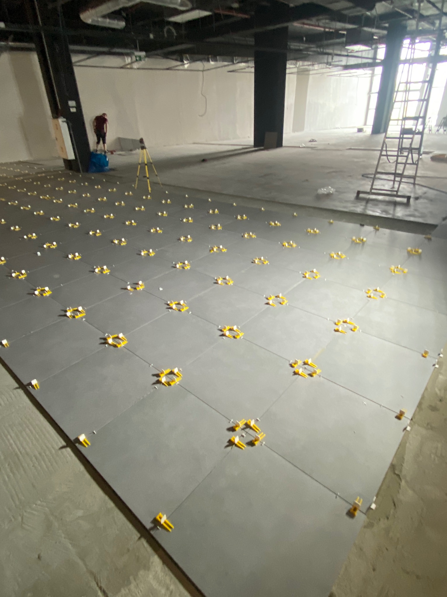 Układanie szarych płytek podłogowych w dużej przestrzeni komercyjnej z użyciem żółtych klinów poziomujących; widoczny laserowy niwelator i drabina w tle.
