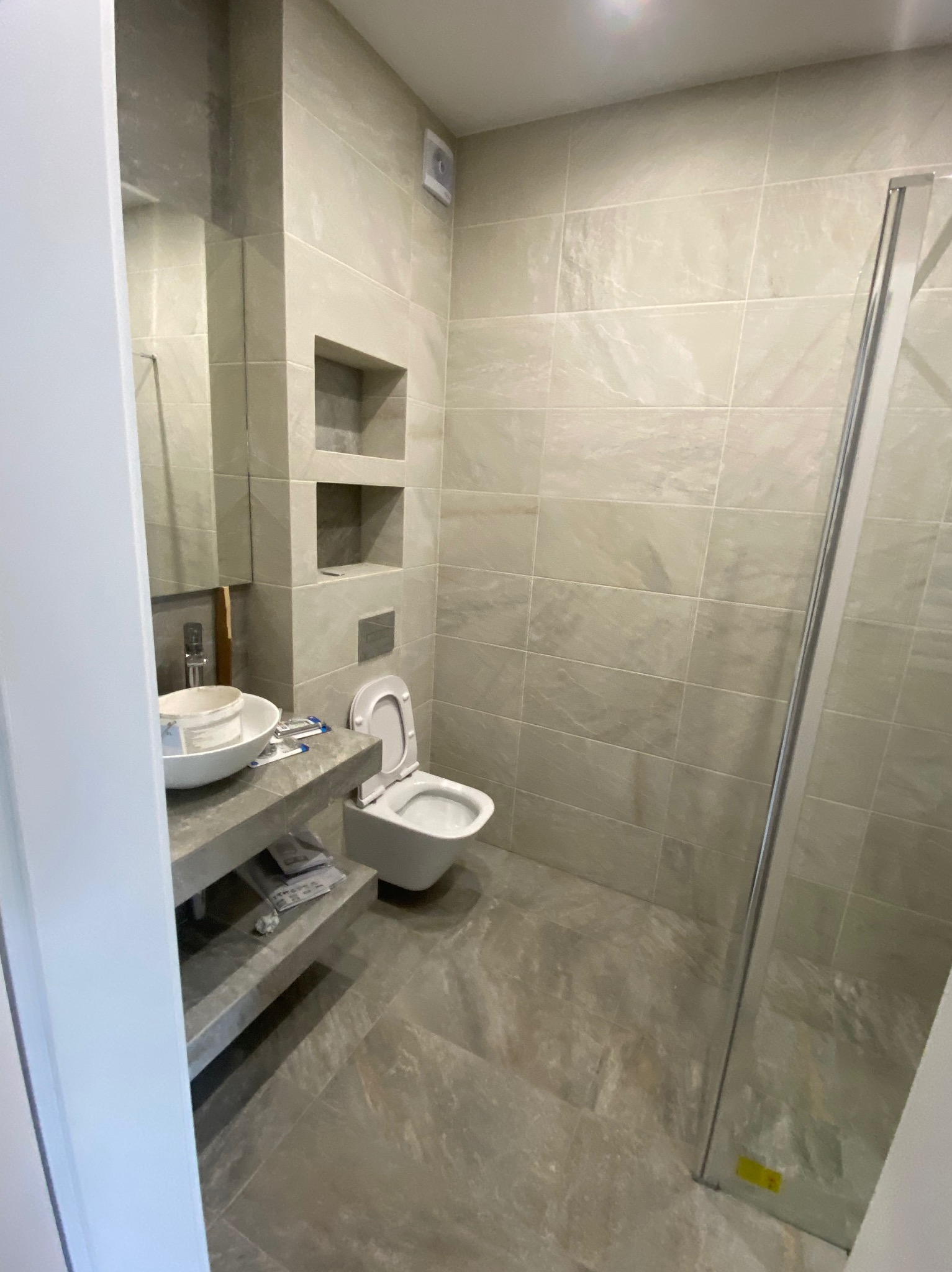 Nowoczesna łazienka w trakcie wykańczania, widoczne płytki imitujące kamień, zabudowa WC, umywalka nablatowa z pozostawionym wiadrem po farbie i narzędziami, kabina prysznicowa z przezroczystego...