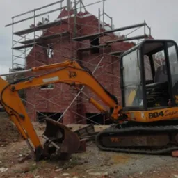 Budowa domu z czerwonej cegły w trakcie wznoszenia murów, widoczne rusztowania oraz koparka JCB 804 Super na pierwszym planie.