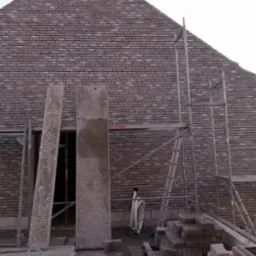 Wznoszenie ściany z cegły klinkierowej, widoczne rusztowanie, ułożone cegły, fragment otworu drzwiowego z betonowymi elementami szalunkowymi.