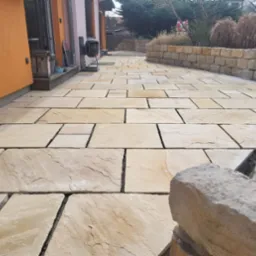 Ułożony z piaskowca taras przed domem z pomarańczową elewacją, widoczny fragment murku oporowego z piaskowca i roślinność w tle.