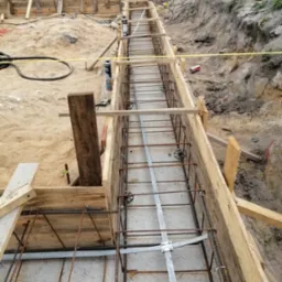 Wykopany rów z drewnianym szalunkiem i zbrojeniem stalowym, przygotowany do zalania betonem, widok z góry na placu budowy.