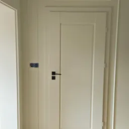 Białe drzwi wewnętrzne z czarną klamką, widoczna instalacja elektryczna w ścianie, parkiet ułożony w jodełkę.