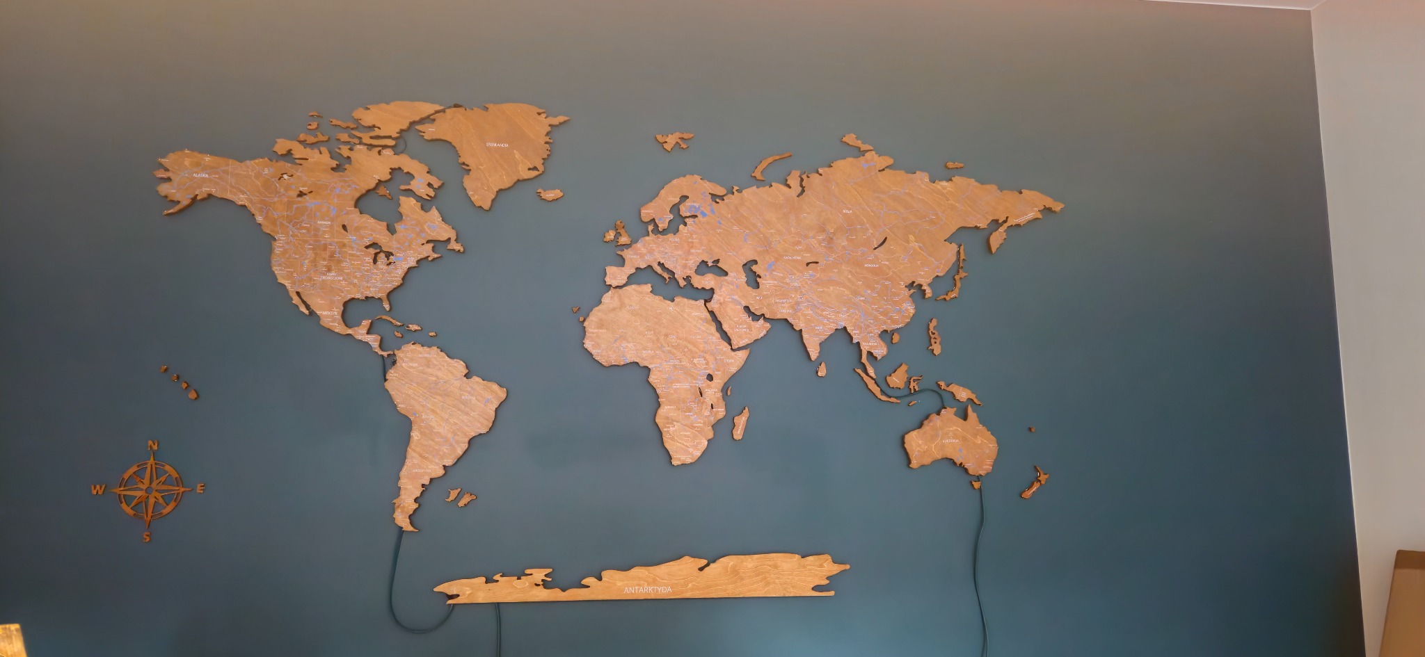 Drewniana mapa świata z podświetleniem LED zamontowana na ścianie pomalowanej na kolor morski, widoczny kompas w lewym dolnym rogu.