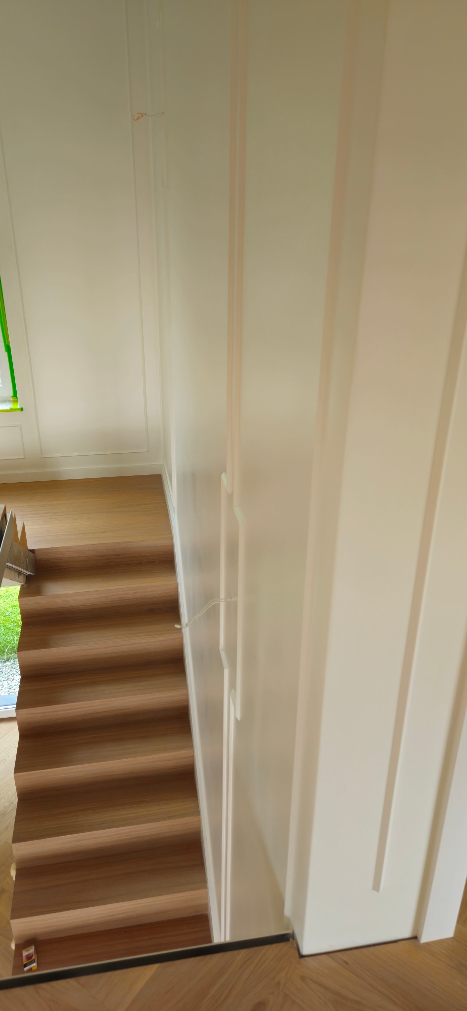 Widok z góry na drewniane schody z jasnymi, panelowymi ścianami i fragmentem okna z zieloną folią ochronną.