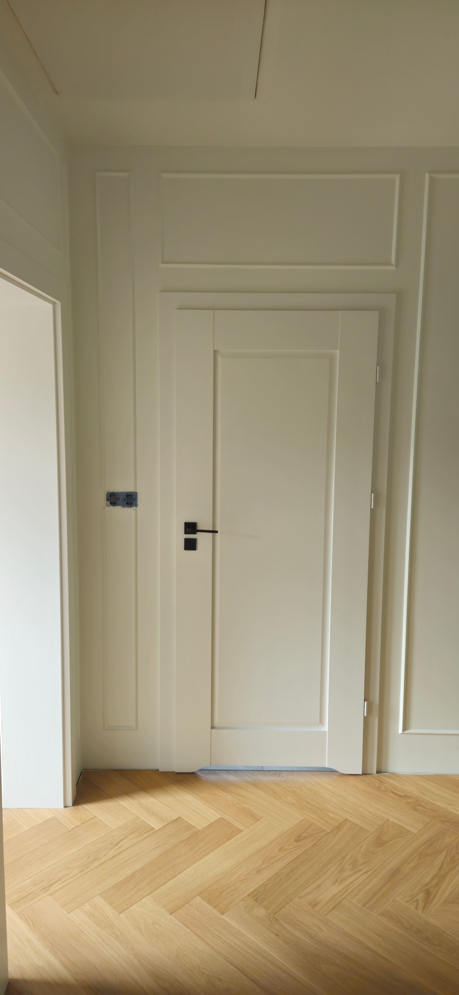 Białe drzwi wewnętrzne z czarną klamką, widoczna instalacja elektryczna w ścianie, parkiet ułożony w jodełkę.