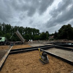 LaSquadra24 Sp. Z O. O. - Przygotowanie podłoża pod budynek: wykop z fundamentami szalunkowymi, ubijarka wibracyjna i wywrotka z piaskiem w tle, pochmurne niebo.
