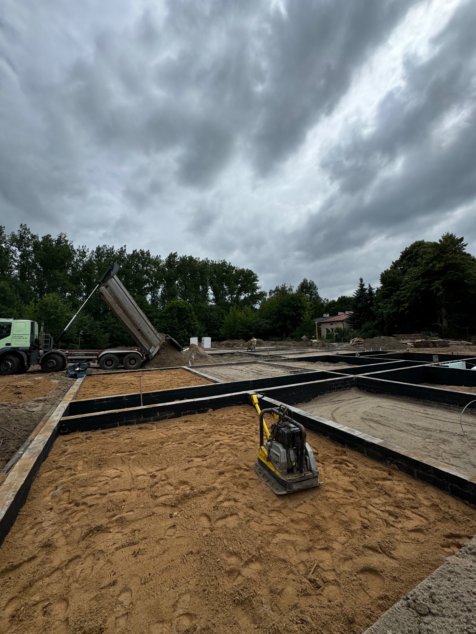 Przygotowanie podłoża pod budynek: wykop z fundamentami szalunkowymi, ubijarka wibracyjna i wywrotka z piaskiem w tle, pochmurne niebo.