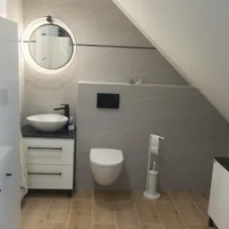 Nowoczesna łazienka na poddaszu z umywalką nablatową, okrągłym lustrem z podświetleniem LED, podwieszaną toaletą i płytkami imitującymi marmur na ścianach oraz drewnopodobnymi płytkami na podłodze.