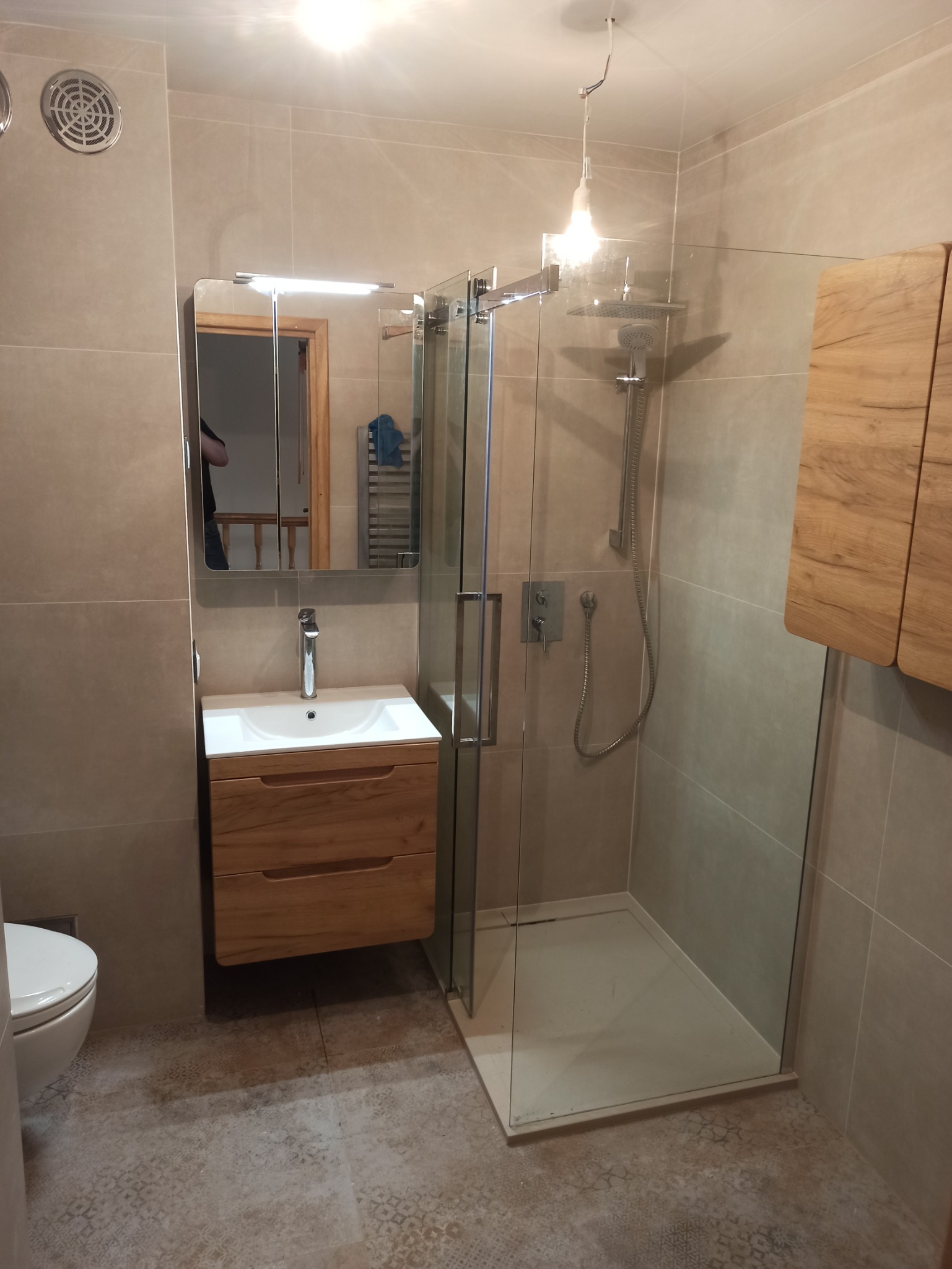 Nowoczesna łazienka z kabiną prysznicową typu walk-in, drewnianą szafką pod umywalką i lustrem z oświetleniem, wykończona beżowymi płytkami i podłogą w delikatny wzór.