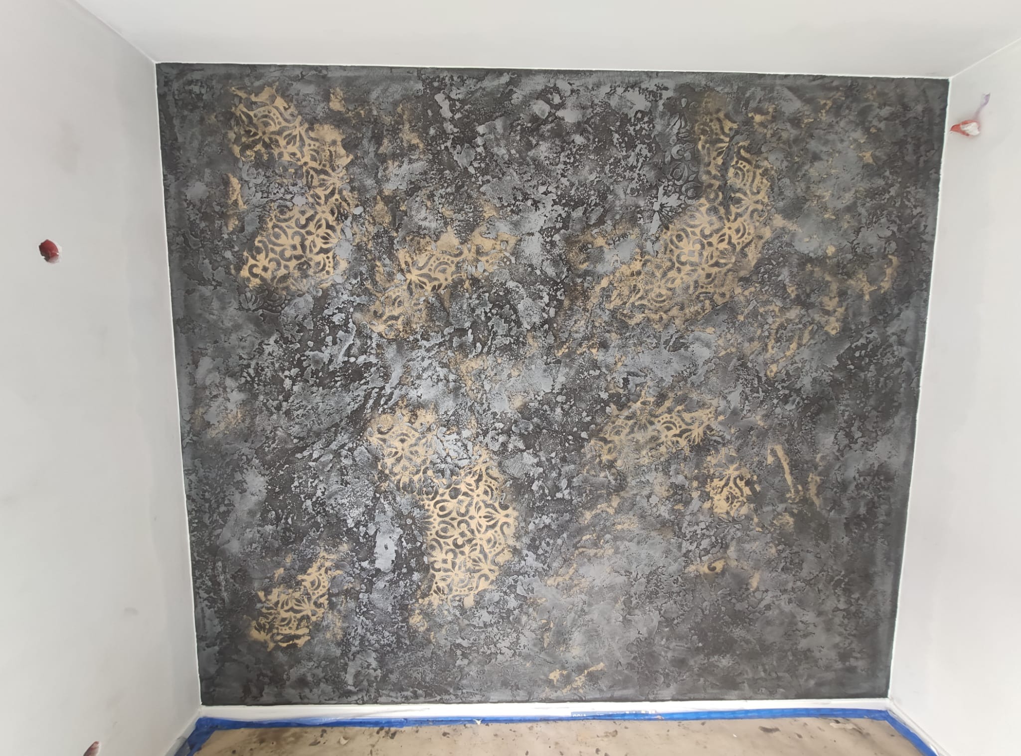 Ściana w pokoju po dekoracyjnym malowaniu w odcieniach szarości i złota z motywem ornamentów, zabezpieczona taśmą malarską przy podłodze.