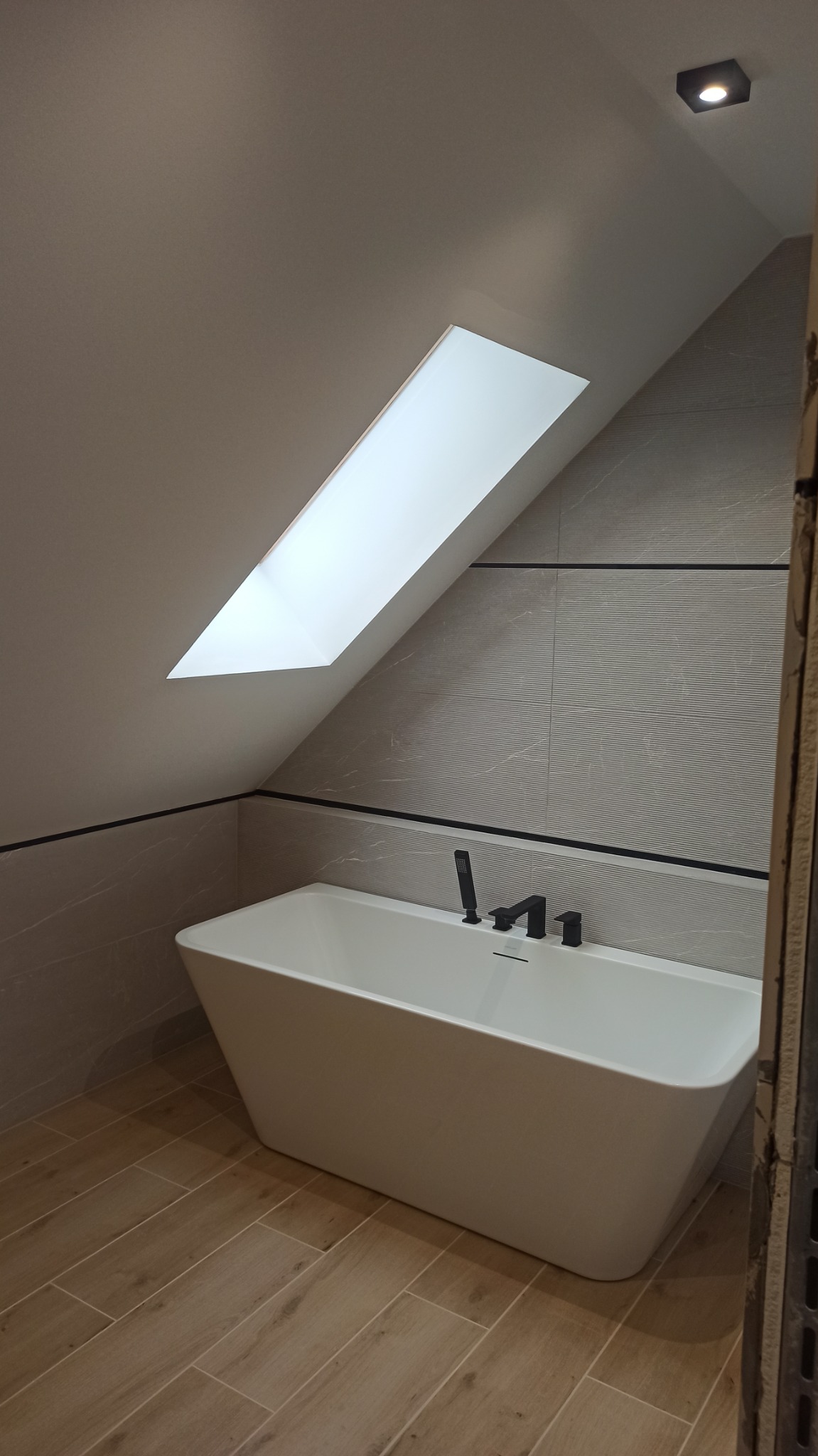 Nowoczesna, minimalistyczna łazienka na poddaszu z wolnostojącą białą wanną, czarną armaturą i oknem dachowym, wykończona płytkami w odcieniach szarości i beżu.