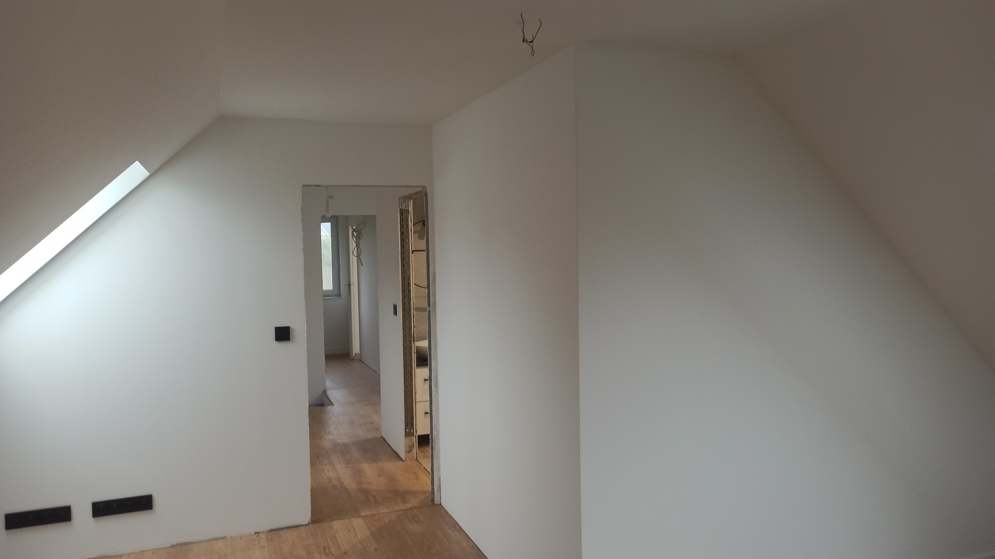 Poddasze po remoncie z oknem dachowym, białe ściany, drewniana podłoga, widoczne gniazdka elektryczne i otwarte drzwi do łazienki z białymi meblami.