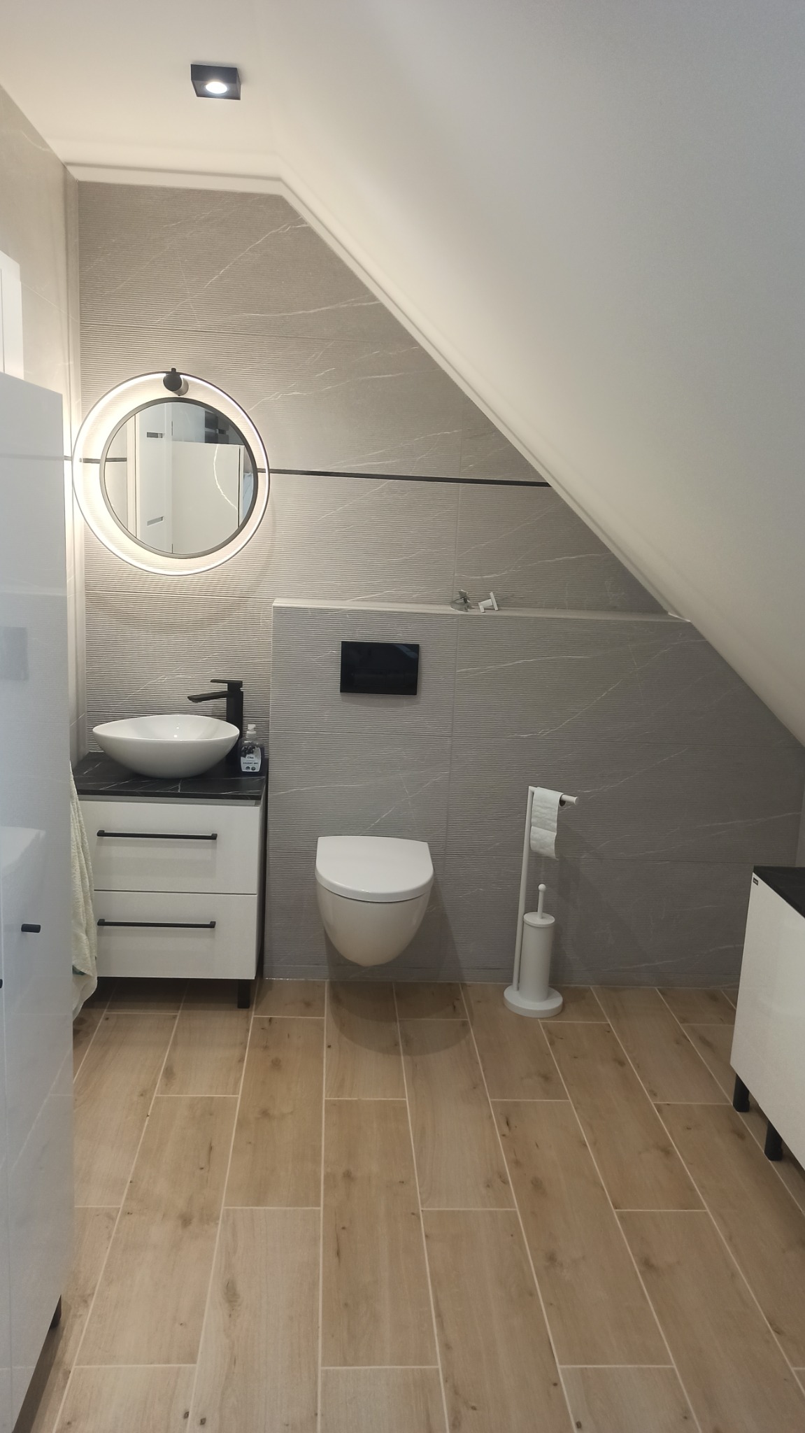 Nowoczesna łazienka na poddaszu z umywalką nablatową, okrągłym lustrem z podświetleniem LED, podwieszaną toaletą i płytkami imitującymi marmur na ścianach oraz drewnopodobnymi płytkami na podłodze.