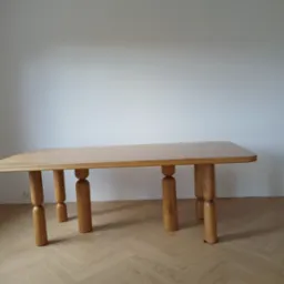 Jasny, dębowy st&oacute;ł z unikalnymi, toczonymi nogami w kształcie walc&oacute;w, ustawiony na podłodze w jodełkę, na tle białej ściany. Minimalistyczny design.