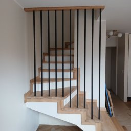 MatWood Usługi Stolarskie - Jasne schody z czarną, minimalistyczną balustradą w nowoczesnym wnętrzu. Widok z dołu, ukazujący detale wykończenia i kontrast kolorystyczny.