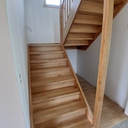 MatWood Usługi Stolarskie - Jasne, drewniane schody z balustradą w minimalistycznym wnętrzu. Widoczne naturalne usłojenie drewna i subtelne oświetlenie z okna na górze.