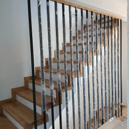 MatWood Usługi Stolarskie - Nowoczesna balustrada z czarnymi, metalowymi słupkami i drewnianym poręczą, zabezpieczona folią ochronną, na tle jasnych, drewnianych schodów.