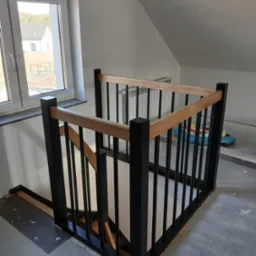 Nowoczesna, czarno-drewniana balustrada schodowa z jasnym drewnem, widok z góry, surowe wykończenie podłogi, duże okno w tle z widokiem na dom.