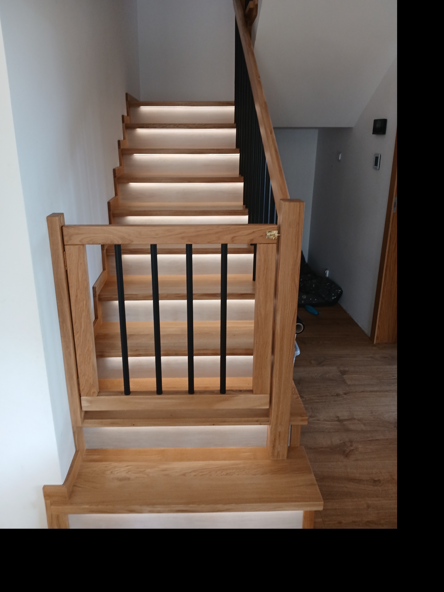 Drewniane schody z podświetleniem LED, z balustradą z czarnymi, pionowymi elementami. Jasne drewno stopni i poręczy kontrastuje z bielą ściany i szarością tła.