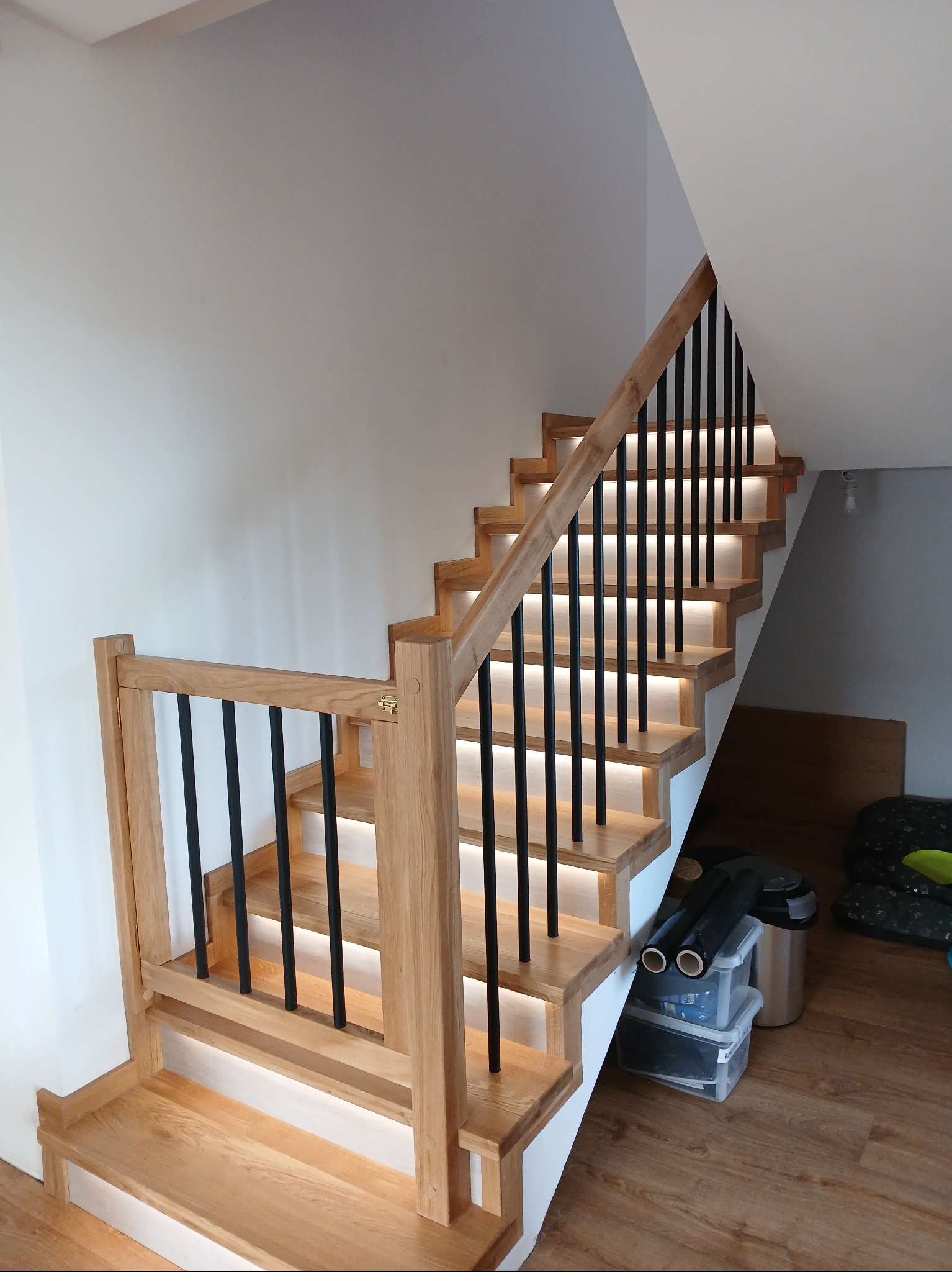 Jasne, dębowe schody z czarną balustradą i podświetleniem LED, zrealizowane w Kołczygłowach. Nowoczesne wykończenie, widok z dołu, detale połączeń elementów.
