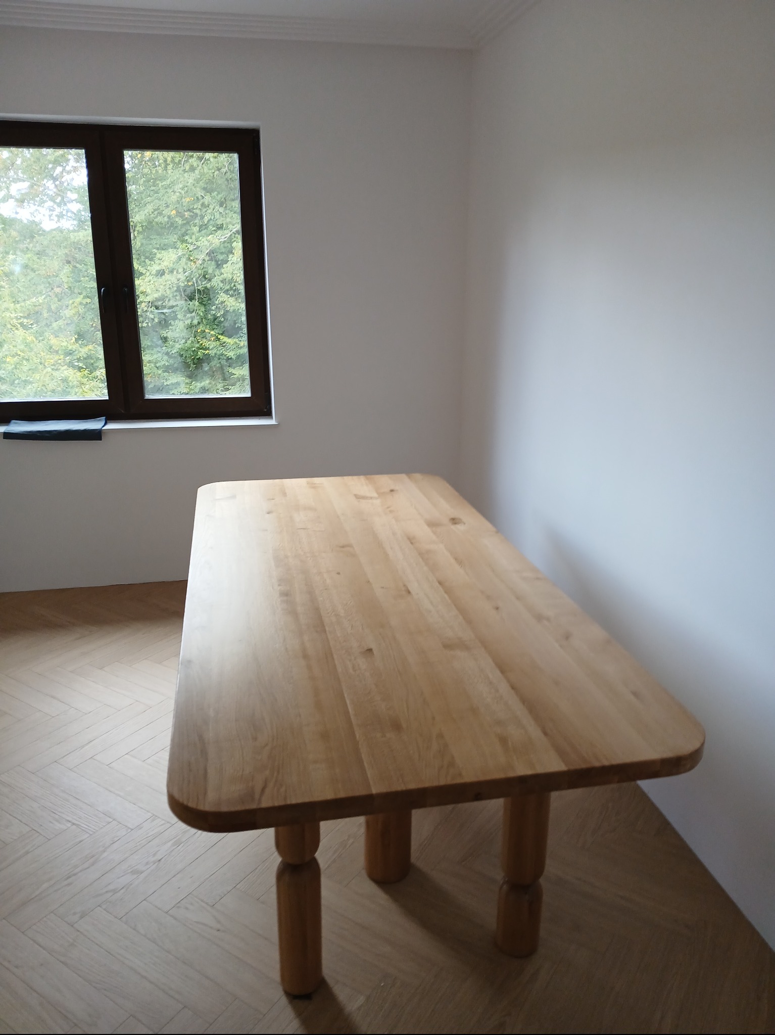 Jasny, drewniany blat stołu z zaokrąglonymi krawędziami i grubymi nogami na tle białej ściany i parkietu, z widokiem na zieleń za oknem.