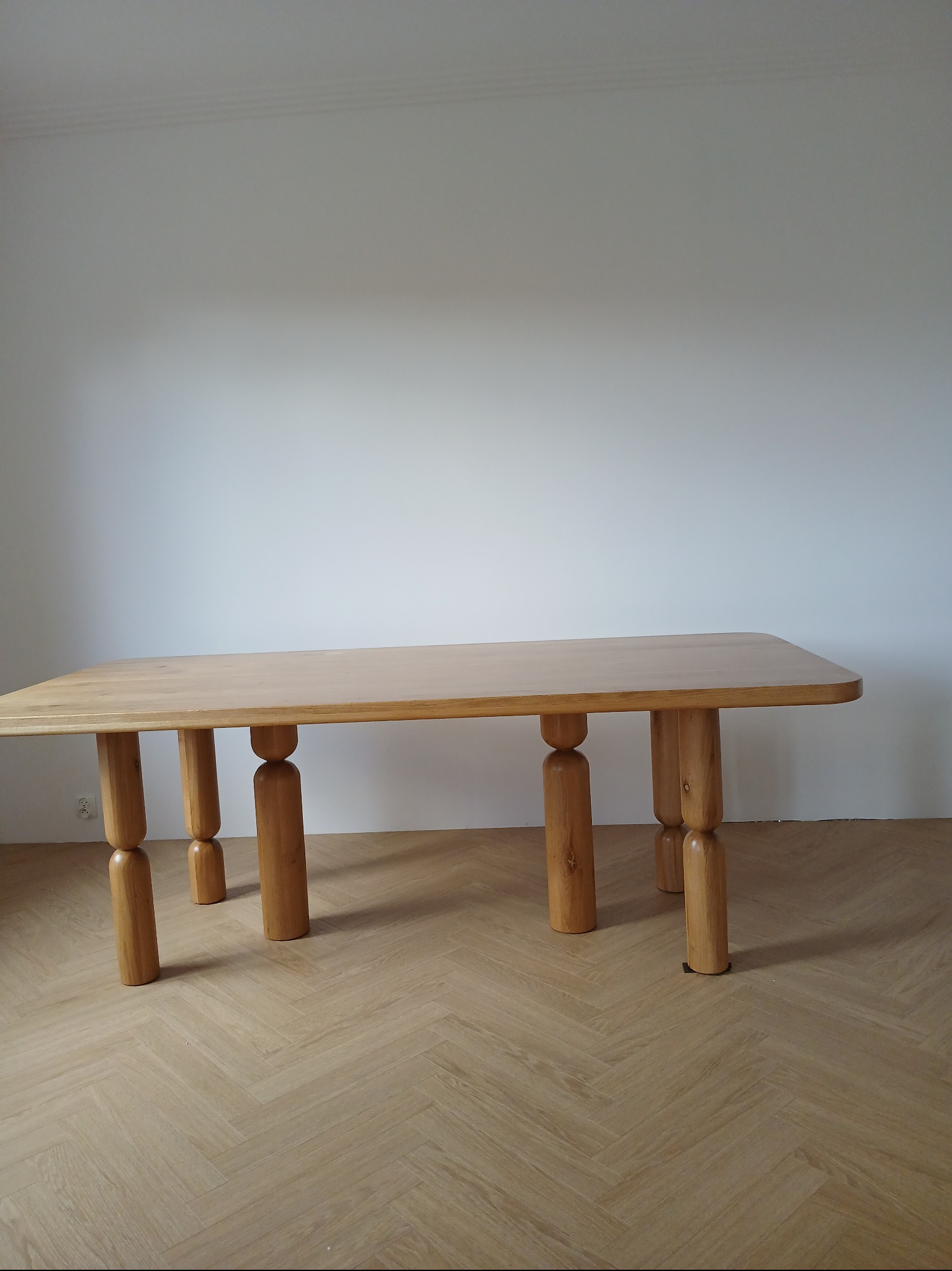 Jasny, dębowy stół z unikalnymi, toczonymi nogami w kształcie walców, ustawiony na podłodze w jodełkę, na tle białej ściany. Minimalistyczny design.