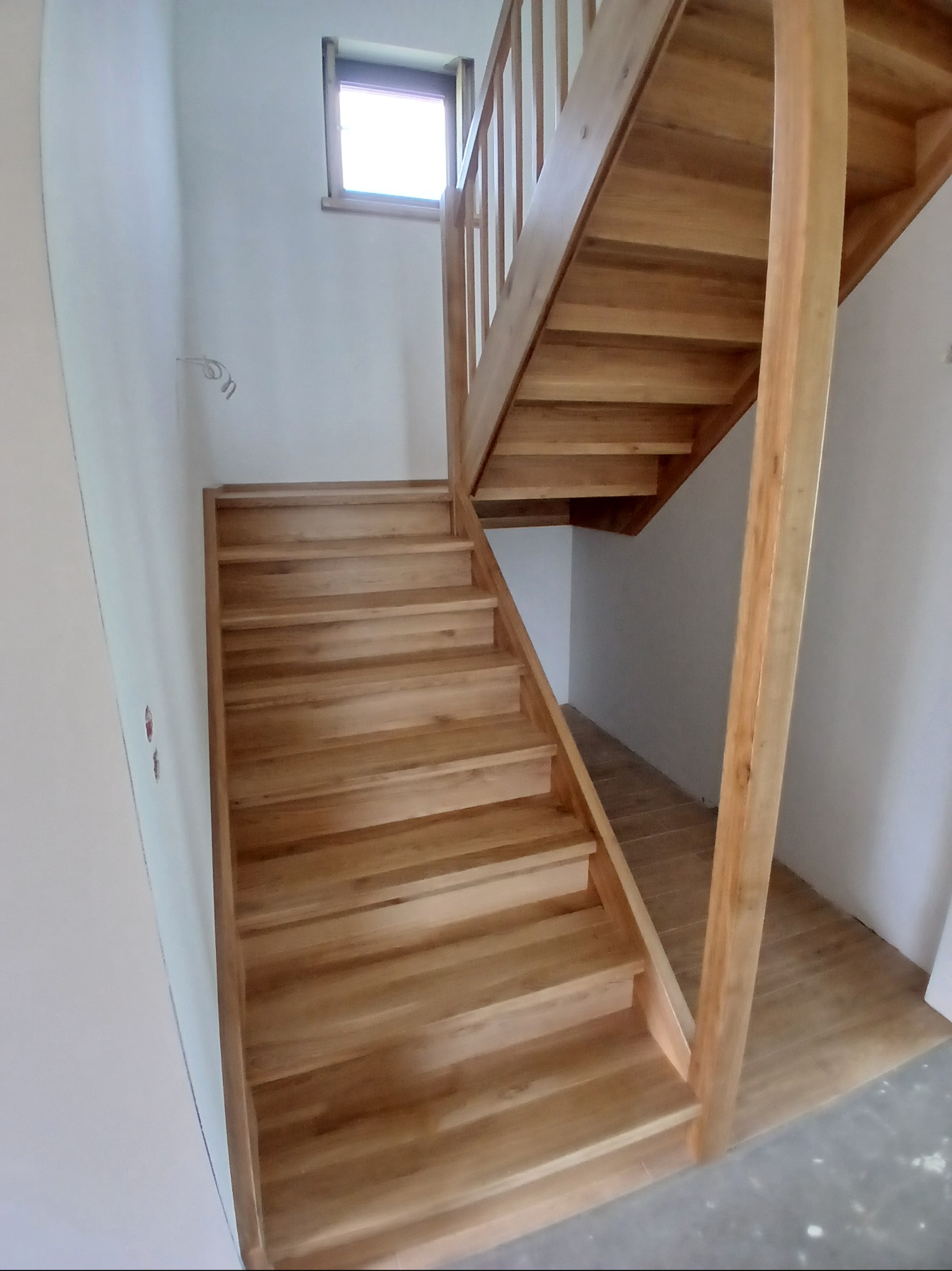 Jasne, drewniane schody z balustradą w minimalistycznym wnętrzu. Widoczne naturalne usłojenie drewna i subtelne oświetlenie z okna na górze.