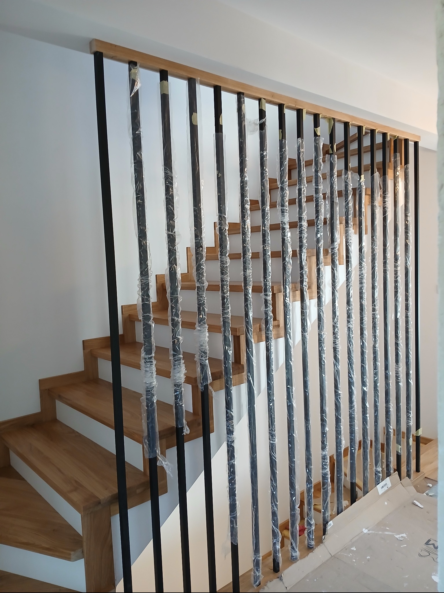Nowoczesna balustrada z czarnymi, metalowymi słupkami i drewnianym poręczą, zabezpieczona folią ochronną, na tle jasnych, drewnianych schodów.