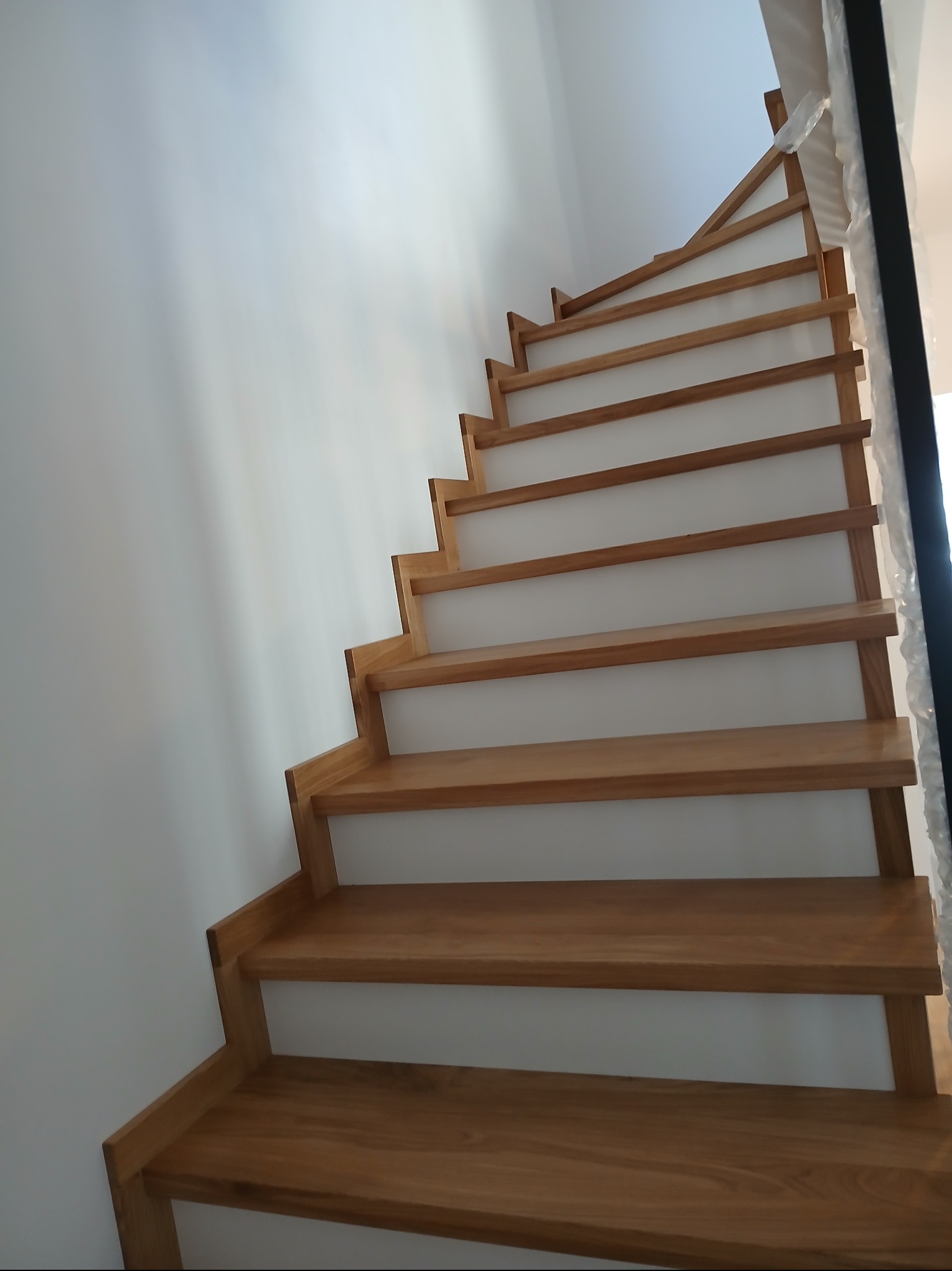 Kątowe ujęcie na jasne, drewniane schody z białymi podstopnicami, w nowoczesnym wnętrzu z białą ścianą. Widoczne zabezpieczenia na balustradzie.