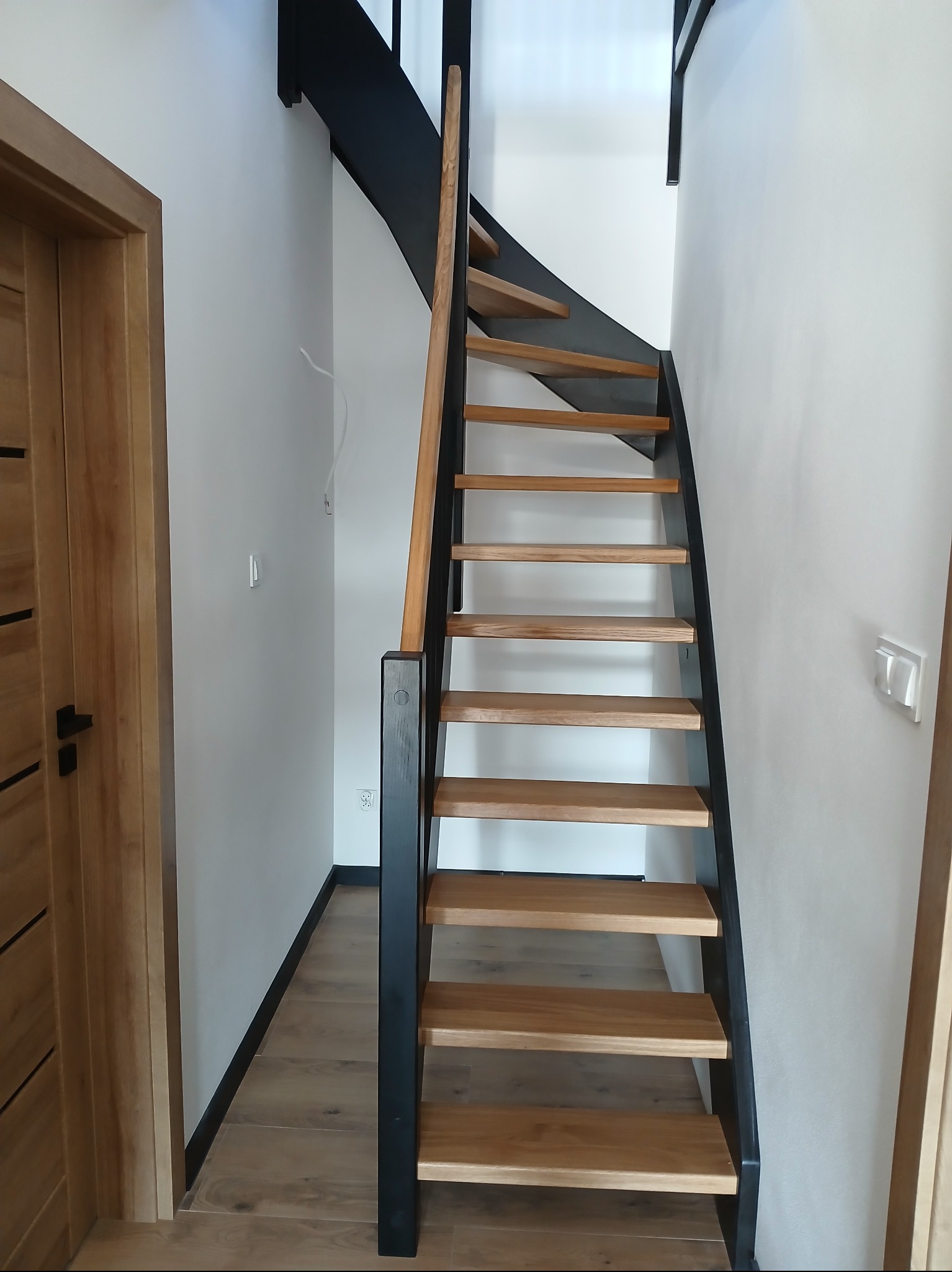 Nowoczesne schody z drewnianymi stopniami i czarną metalową konstrukcją, widoczne z dołu, z fragmentem drewnianych drzwi po lewej stronie.