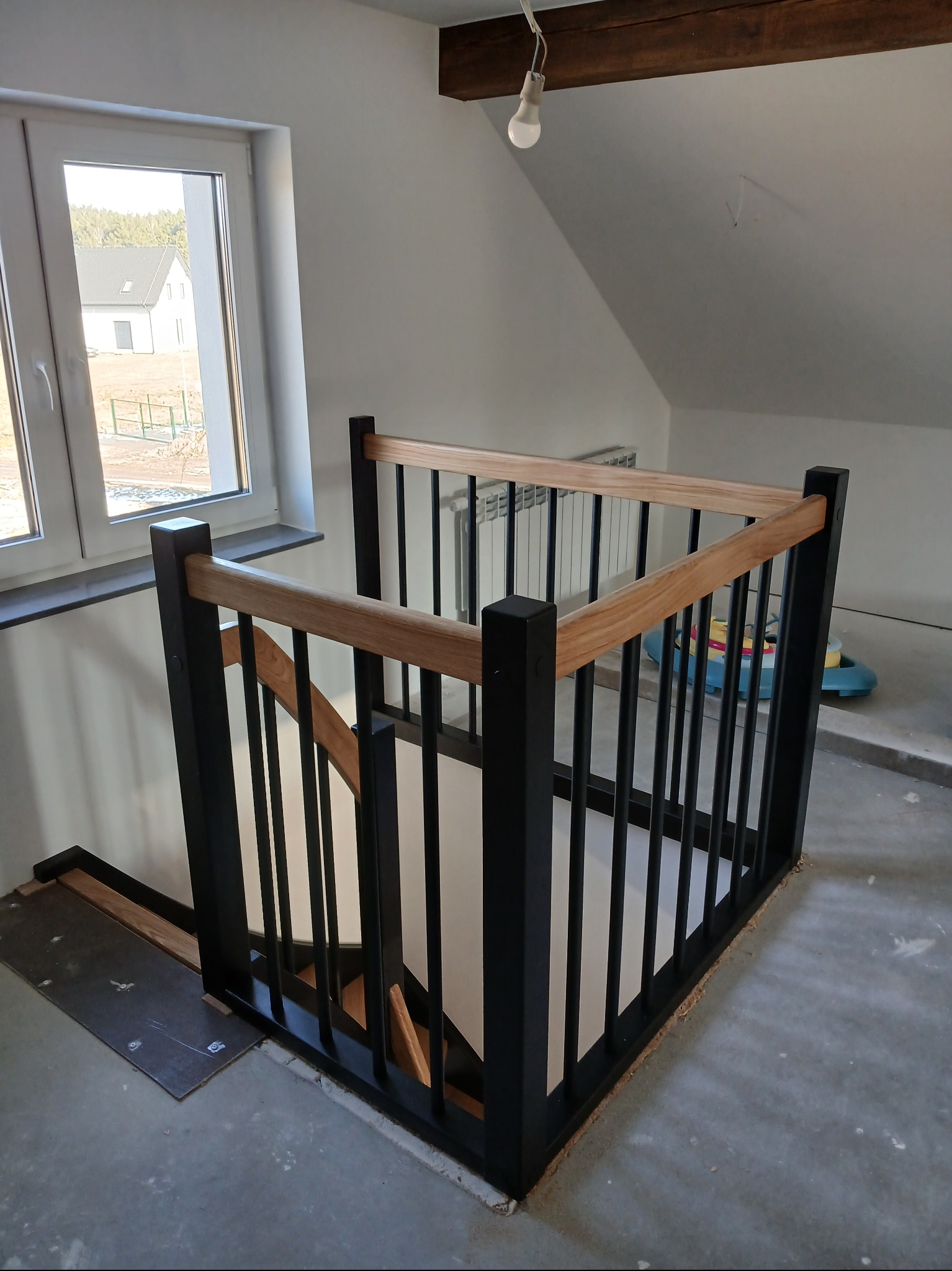 Nowoczesna, czarno-drewniana balustrada schodowa z jasnym drewnem, widok z góry, surowe wykończenie podłogi, duże okno w tle z widokiem na dom.
