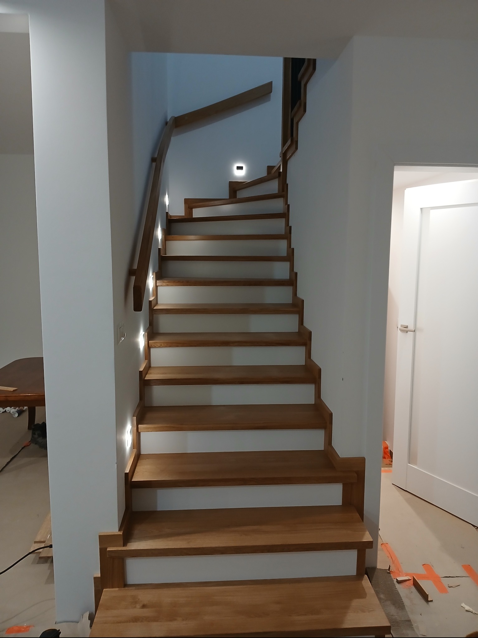 Nowoczesne schody z drewna z oświetleniem LED, prowadzące na górę. Widoczne detale wykończenia i minimalistyczny design. Białe ściany, dębowe stopnie i poręcz.