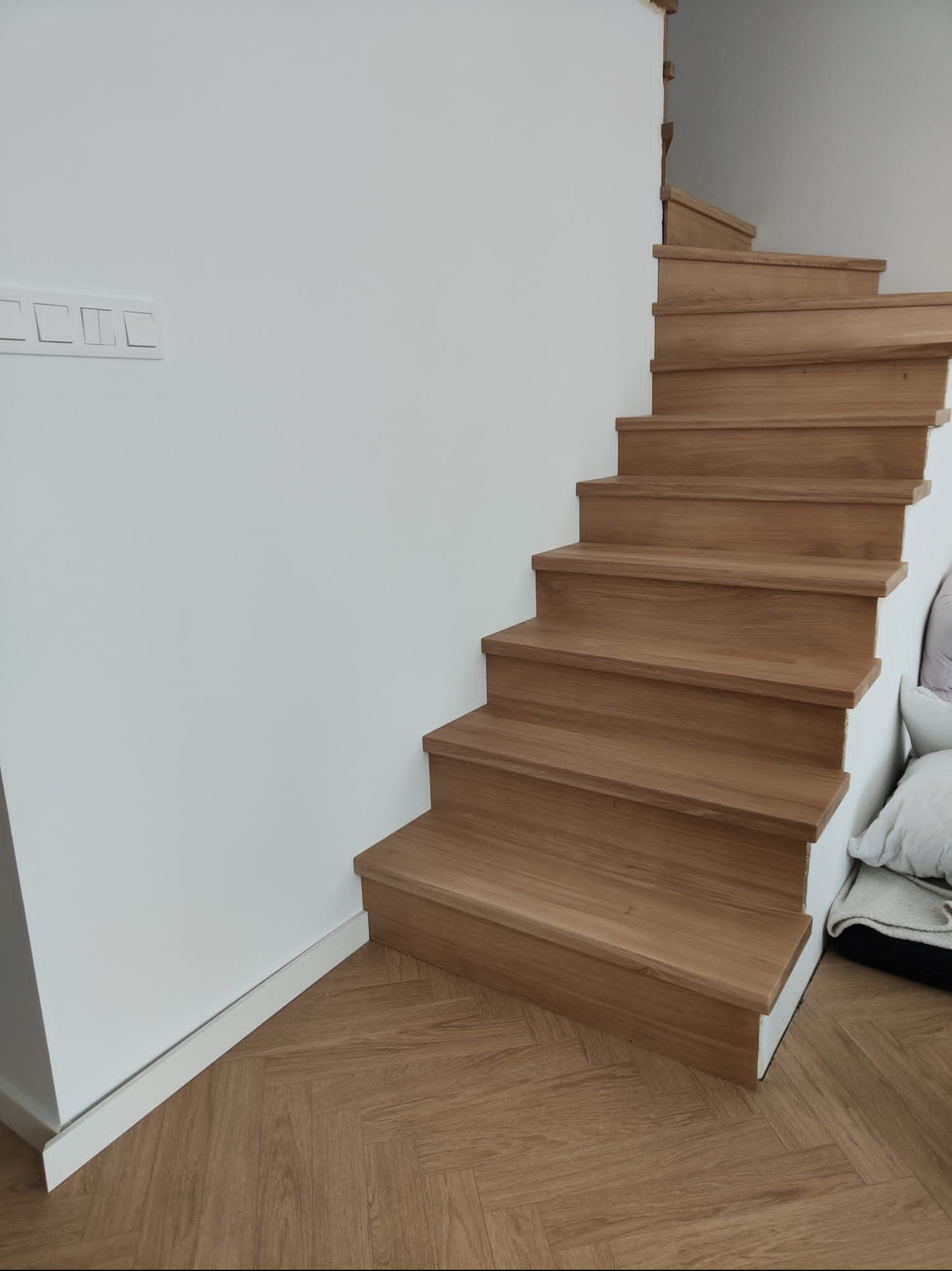 Jasne, dębowe schody z widocznym wzorem parkietu jodełkowego na podłodze. Minimalistyczne wykończenie ścian i schodów podkreśla naturalny charakter drewna.